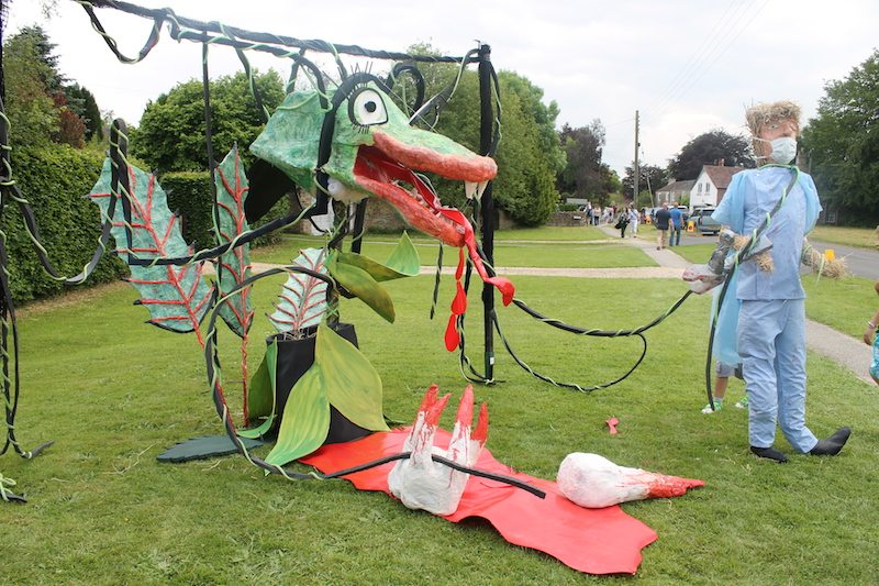 A dragon scarecrow display on the village green at the Scarecrow Festival, with people on the pavement behind